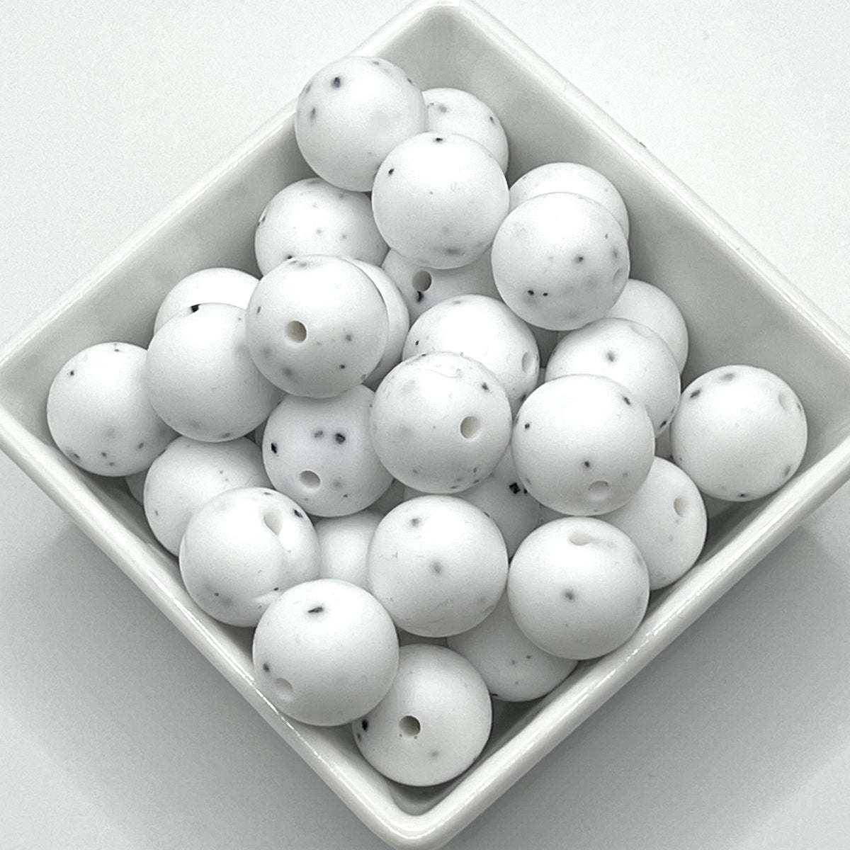 White round craft beads with small black speckles and pre-drilled holes, displayed in a square white dish on a light gray background.