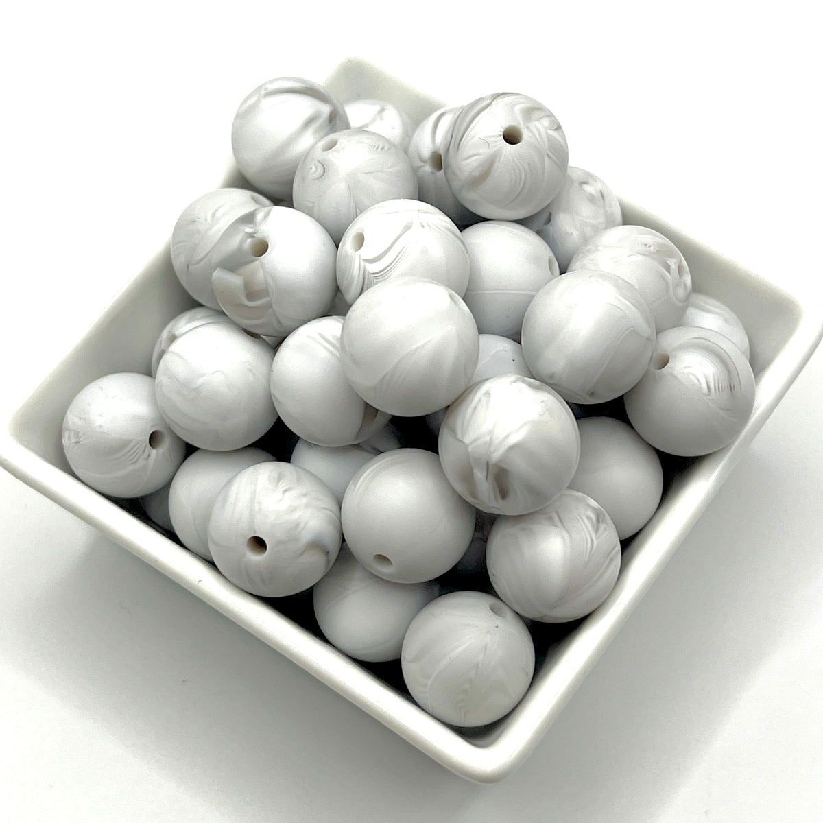 A pile of round, glossy white beads with a marbled swirl pattern, each featuring a small hole, displayed in a small white square dish on a white background.